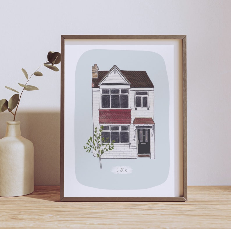 Op de afbeelding: Een ingelijste illustratie van een huis met twee verdiepingen met een rode bakstenen gevel, zwarte ramen en een zwarte deur. Het huis heeft een kleine boom ervoor en de tekst "J&B" eronder.