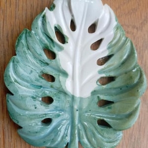 K&ouml;nnte beinhalten: Eine blattf&ouml;rmige Ablageschale mit Monstera-Design und ausgestanzten L&ouml;chern. Die Schale ist zweifarbig gestaltet, wobei eine Seite in schlichtem Wei&szlig; und die andere Seite in einem strukturierten, gesprenkelten Salbeigr&uuml;n gehalten ist.