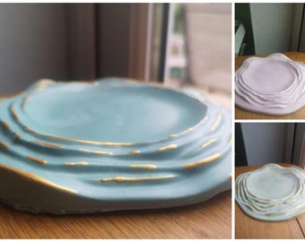 Könnte beinhalten: Ein kreisförmiges Tablett mit wellenförmigem Randdesign. Das Tablett verfügt über eine glatte Mitte und erhabene konzentrische Ringe am Rand, die mit metallischer Goldfarbe auf den Kanten akzentuiert sind. Erhältlich in Hellblau, Blassrosa und Weiß.