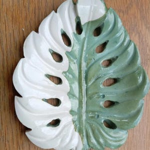 K&ouml;nnte beinhalten: Eine Keramikschale in Form eines Monstera-Blattes mit zweifarbigem Design. Die linke Seite ist mit einer gl&auml;nzenden wei&szlig;en Glasur versehen, w&auml;hrend die rechte Seite eine gedeckte salbeigr&uuml;ne Glasur aufweist. Das Blatt hat Aussparungen.