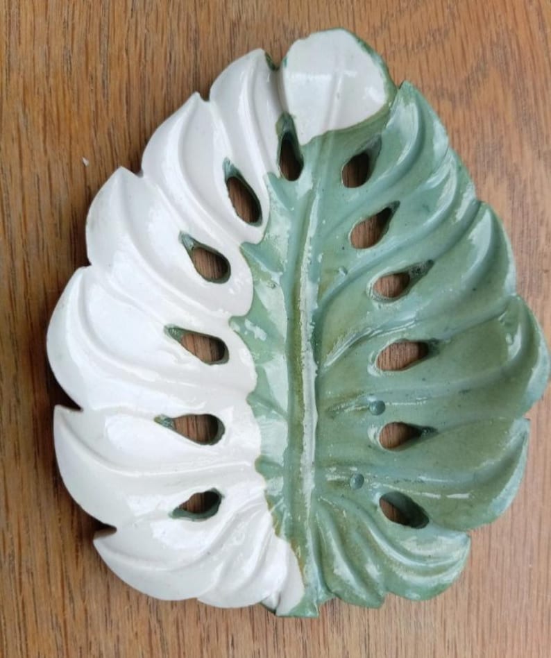 K&ouml;nnte beinhalten: Eine Keramikschale in Form eines Monstera-Blattes mit zweifarbigem Design. Die linke Seite ist mit einer gl&auml;nzenden wei&szlig;en Glasur versehen, w&auml;hrend die rechte Seite eine gedeckte salbeigr&uuml;ne Glasur aufweist. Das Blatt hat Aussparungen.