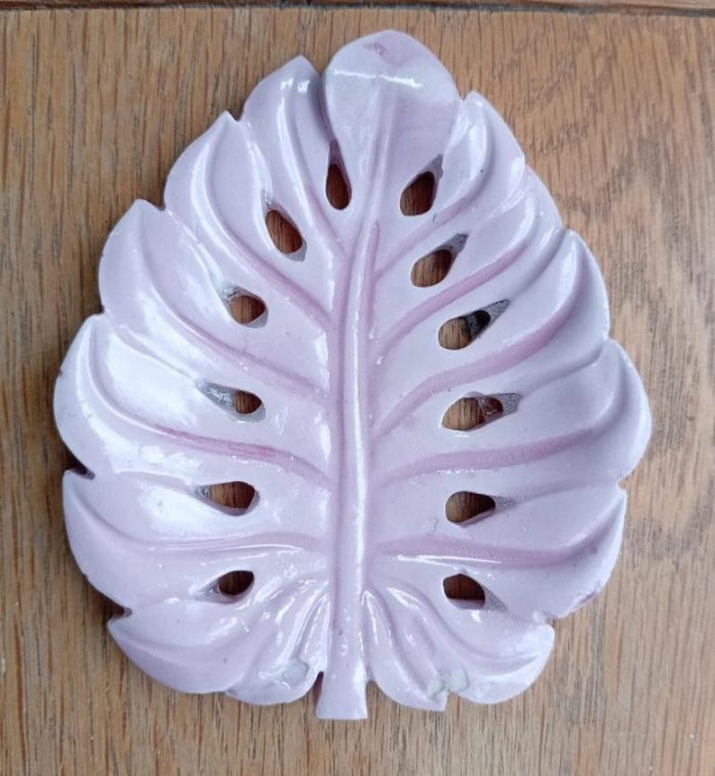 K&ouml;nnte beinhalten: Eine hellrosa, blattf&ouml;rmige Schale mit einer zentralen Blattader und mehreren ausgestanzten &Ouml;ffnungen an den Seiten, die dem Aussehen eines Monstera-Blattes nachempfunden ist. Das Objekt hat eine glatte, gl&auml;nzende Oberfl&auml;che und eine definierte, symmetrische Struktur.