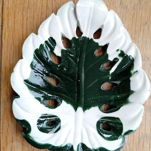 K&ouml;nnte beinhalten: Eine Keramikschale in Form eines Monstera-Blattes mit gl&auml;nzender Oberfl&auml;che. Das Blatt hat ein zweifarbiges Design mit einem tiefen waldgr&uuml;nen Zentrum und wei&szlig;en R&auml;ndern sowie mehreren ausgestanzten L&ouml;chern in der Blattstruktur.