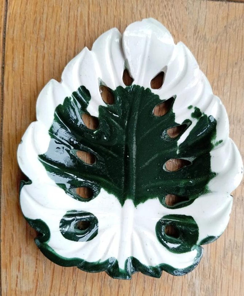 K&ouml;nnte beinhalten: Eine Keramikschale in Form eines Monstera-Blattes mit gl&auml;nzender Oberfl&auml;che. Das Blatt hat ein zweifarbiges Design mit einem tiefen waldgr&uuml;nen Zentrum und wei&szlig;en R&auml;ndern sowie mehreren ausgestanzten L&ouml;chern in der Blattstruktur.