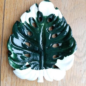 K&ouml;nnte beinhalten: Eine gl&auml;nzende Keramikschale in Form eines Monstera-Blattes mit einem panaschierten Muster aus tiefem Waldgr&uuml;n und reinem Wei&szlig;. Das Blattdesign weist charakteristische Ausschnitte und Blattadern sowie eine glatte Oberfl&auml;che auf.