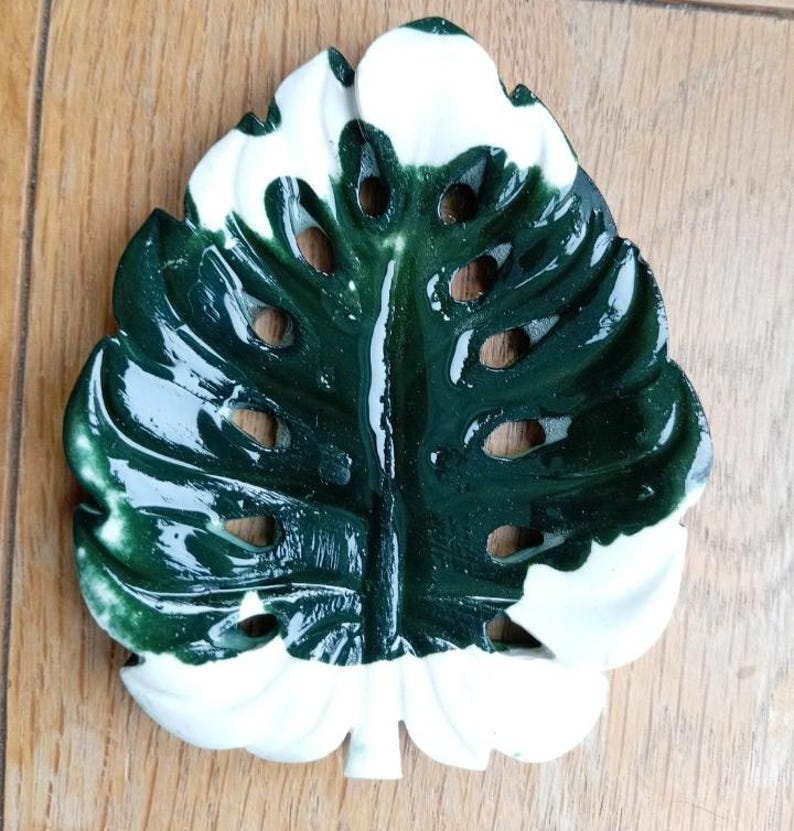 K&ouml;nnte beinhalten: Eine gl&auml;nzende Keramikschale in Form eines Monstera-Blattes mit einem panaschierten Muster aus tiefem Waldgr&uuml;n und reinem Wei&szlig;. Das Blattdesign weist charakteristische Ausschnitte und Blattadern sowie eine glatte Oberfl&auml;che auf.