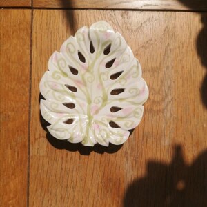 K&ouml;nnte beinhalten: Eine wei&szlig;e Keramikschale in Form eines Monstera-Blattes mit ausgeschnittenen Bereichen. Die Oberfl&auml;che ist mit handgemalten hellgr&uuml;nen, geschwungenen Rankenmustern und kleinen, verstreuten rosa Punkten auf dem glatten, wei&szlig;en Finish verziert.