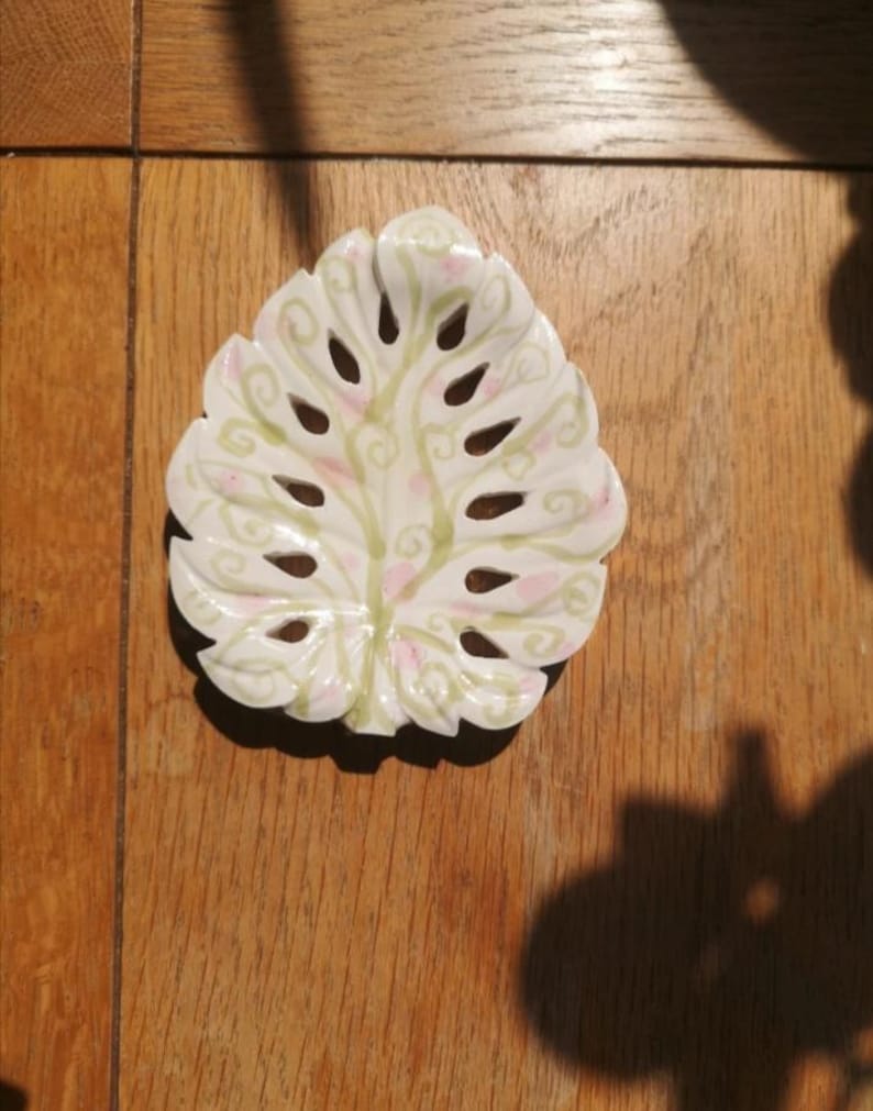 K&ouml;nnte beinhalten: Eine wei&szlig;e Keramikschale in Form eines Monstera-Blattes mit ausgeschnittenen Bereichen. Die Oberfl&auml;che ist mit handgemalten hellgr&uuml;nen, geschwungenen Rankenmustern und kleinen, verstreuten rosa Punkten auf dem glatten, wei&szlig;en Finish verziert.