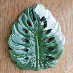 K&ouml;nnte beinhalten: Eine gl&auml;nzende Keramikschale in Form eines Monstera-Blattes. Das Blatt ist gr&ouml;&szlig;tenteils in einem tiefen Waldgr&uuml;n glasiert, mit einem deutlich abgesetzten wei&szlig;en Bereich im oberen rechten Teil. Es weist mehrere Ausstanzungen auf.