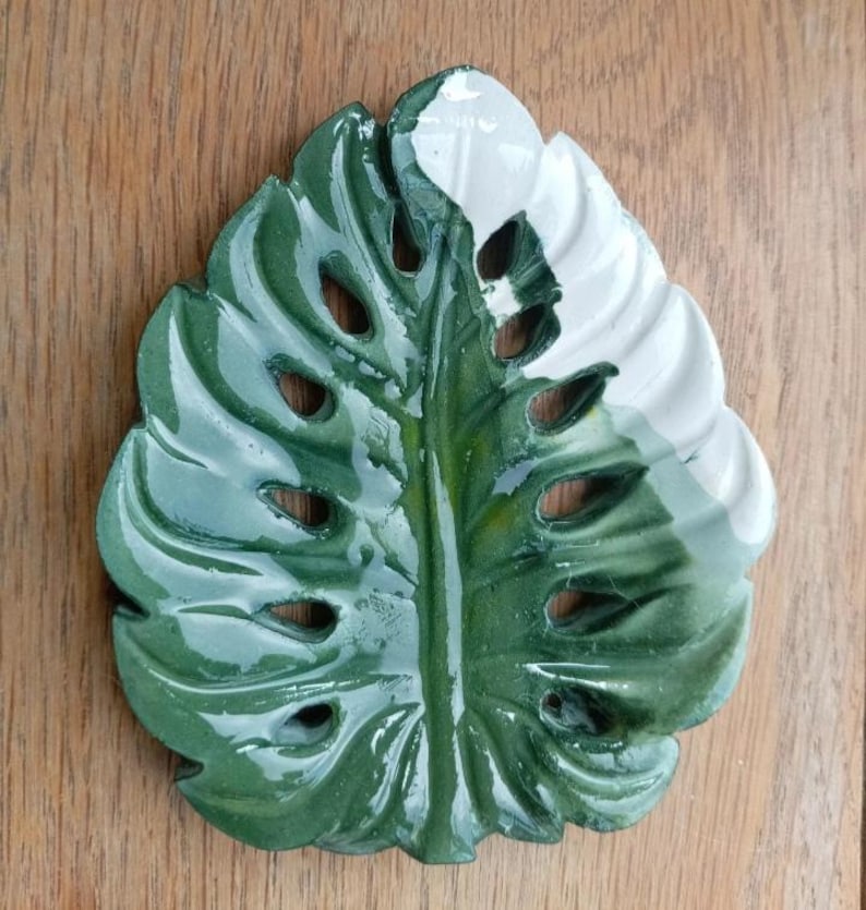K&ouml;nnte beinhalten: Eine gl&auml;nzende Keramikschale in Form eines Monstera-Blattes. Das Blatt ist gr&ouml;&szlig;tenteils in einem tiefen Waldgr&uuml;n glasiert, mit einem deutlich abgesetzten wei&szlig;en Bereich im oberen rechten Teil. Es weist mehrere Ausstanzungen auf.