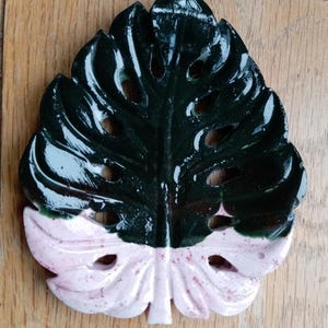 K&ouml;nnte beinhalten: Eine blattf&ouml;rmige Schale mit gl&auml;nzender Oberfl&auml;che, die in einen dunkelgr&uuml;nen oberen und einen hellrosa unteren Bereich unterteilt ist. Das Objekt weist durchgehende Aussparungen auf, die einem Monstera-Blatt nachempfunden sind.