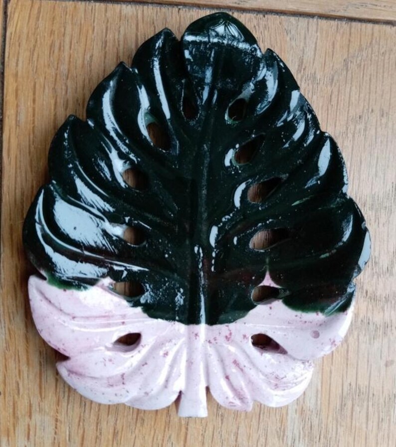 K&ouml;nnte beinhalten: Eine blattf&ouml;rmige Schale mit gl&auml;nzender Oberfl&auml;che, die in einen dunkelgr&uuml;nen oberen und einen hellrosa unteren Bereich unterteilt ist. Das Objekt weist durchgehende Aussparungen auf, die einem Monstera-Blatt nachempfunden sind.