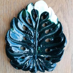 K&ouml;nnte beinhalten: Eine gl&auml;nzende, dunkelgr&uuml;ne Schale in Form eines Monstera-Blattes mit einem markanten wei&szlig;en Bereich an der Spitze. Das Keramikobjekt hat eine glatte, reflektierende Oberfl&auml;che mit ausgestanzten L&ouml;chern, die die Blattstruktur nachahmen.