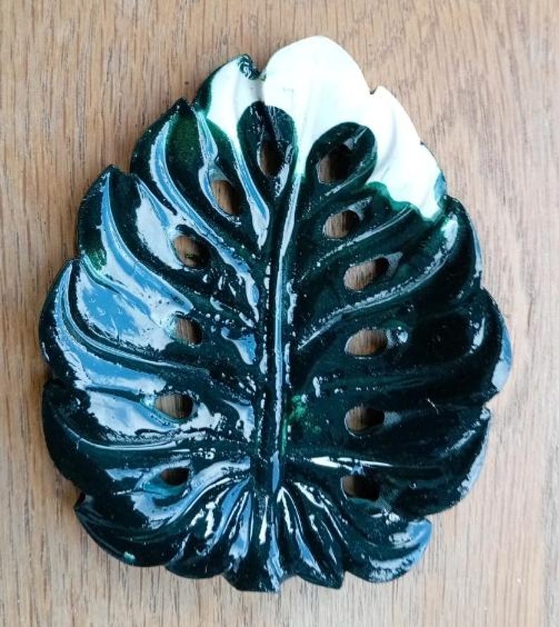 K&ouml;nnte beinhalten: Eine gl&auml;nzende, dunkelgr&uuml;ne Schale in Form eines Monstera-Blattes mit einem markanten wei&szlig;en Bereich an der Spitze. Das Keramikobjekt hat eine glatte, reflektierende Oberfl&auml;che mit ausgestanzten L&ouml;chern, die die Blattstruktur nachahmen.