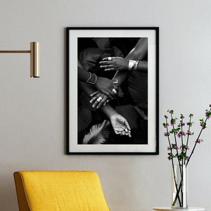 This is an environmental shot of the print in a home. A yellow chair near a wall where the print hangs. The image contains 4 black women's hands reaching and clasping each other, adorned in silver jewelry.