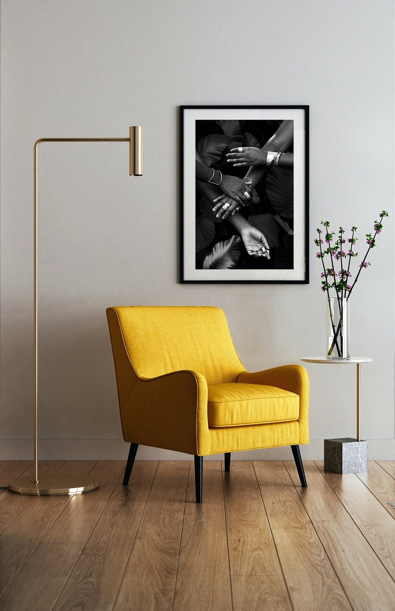 This is an environmental shot of the print in a home. A yellow chair near a wall where the print hangs. The image contains 4 black women's hands reaching and clasping each other, adorned in silver jewelry.