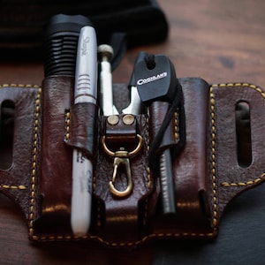 May include: A brown leather multi-tool organiser with yellow stitching. It holds a flashlight, a Sharpie marker, a screwdriver, and a Coghlan's fire starter. The organiser has a brass-coloured clasp.