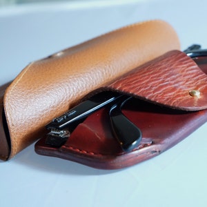 May include: Black-framed spectacles are placed inside a red leather case, alongside a tan leather glasses case. The cases are open, displaying the glasses. The cases are made of textured leather.