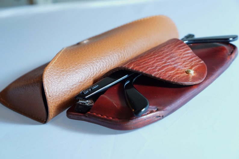 May include: Black-framed spectacles are placed inside a red leather case, alongside a tan leather glasses case. The cases are open, displaying the glasses. The cases are made of textured leather.