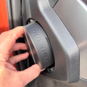 Può includere: Un componente automobilistico nero e circolare con la scritta "4Runner" in rilievo sul lato viene installato in un pannello interno grigio scuro. Il componente è tenuto da una mano, con una portiera arancione sullo sfondo.