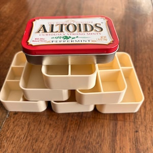 Puede incluir: Una lata de Altoids roja y plateada se encuentra encima de una pila de recipientes de almacenamiento beige compartimentados. La lata dice "Altoids, Curiously Strong Mints, Peppermint" con un peso neto de 50g. Los recipientes están diseñados para organizar objetos pequeños.