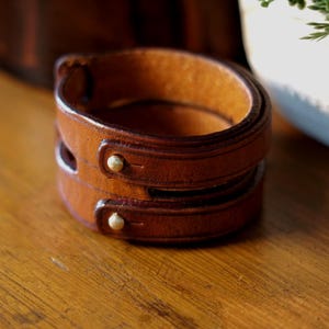May include: A brown leather cuff bracelet with two layers, each secured with a gold-colored stud. The bracelet is made of a rich, textured leather and sits on a wooden surface.