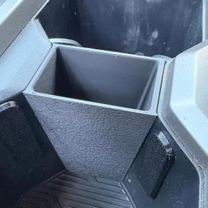 May include: A black, textured, rectangular storage container, possibly for a vehicle, is shown. The container is open at the top and has a textured exterior. It is surrounded by other vehicle components, including a floor mat.