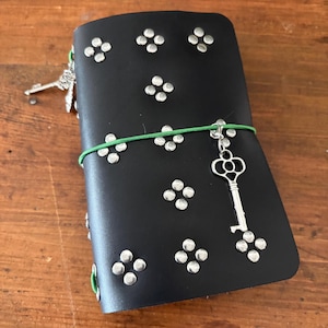 May include: A black journal cover featuring a pattern of silver-toned metal studs arranged in groups of four. A bright green elastic band wraps around the center, holding a silver-toned skeleton key charm. Additional silver charms hang from the side.