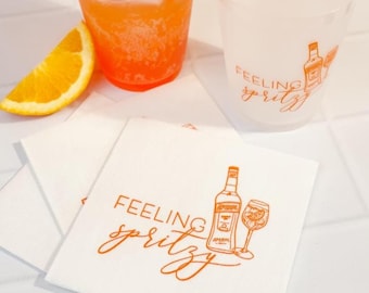 May include: White paper napkins featuring an orange graphic of a bottle, a stemmed glass, and the text Feeling spritzy. Beside them are a translucent plastic cup with the same design, an orange cocktail with ice, and an orange slice.