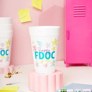 May include: White cup with floral design and the text "happy FDOC" in teal and purple. The cup is on a pink stand, with a pink locker in the background. Other cups and office supplies are also visible.