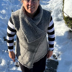 May include: A woman wearing a grey knitted vest with a black and white striped long-sleeved shirt underneath. She is wearing black trousers and is standing in the snow.