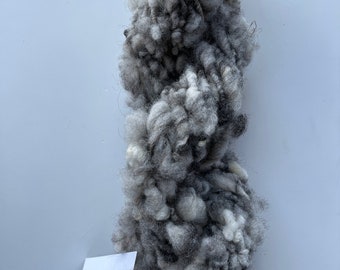 Puede incluir: Un ovillo de hilo grueso en tonos grises y blancos. El hilo está teñido a mano y tiene un aspecto esponjoso y texturizado. La etiqueta dice "Hilo teñido a mano, nube gris tormenta, 100% lana, 100g, 100m".