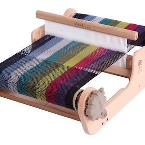 May include: A wooden loom with a colourful woven fabric on it. The loom has two wooden spools on either side and a wooden shuttle in the middle. The fabric is woven with a variety of colours, including red, green, blue, and yellow.