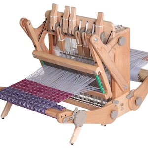 May include: A wooden loom with a partially woven fabric in shades of purple, red, and white. The loom has a wooden frame with a metal warp and a set of heddles. The loom is set up for weaving a tapestry or other fabric.