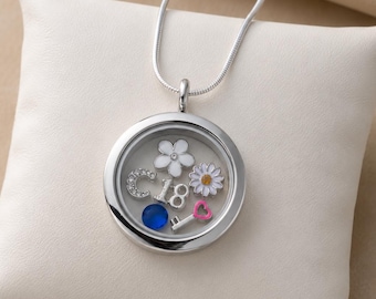Puede incluir: Un collar con colgante circular plateado con frente de cristal transparente que contiene dijes flotantes: una flor blanca, una margarita, una letra C con cristales, el número 18, una cuenta azul y una llave plateada con un corazón rosa.