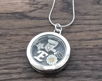 Puede incluir: Collar con un dije de medallón plateado y un frente de vidrio. El medallón contiene un gorro de graduación, un rollo, una estrella, una flor y la letra "E".