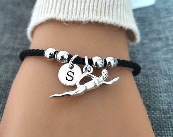 Puede incluir: Un brazalete de cordón negro con cuentas plateadas y un dije de nadador. El dije tiene un pequeño disco plateado con la letra "S" en él.