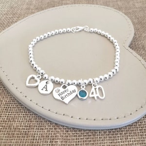 May include: Silver charm bracelet with a heart charm, a charm with the letter A, a heart charm with the words "Happy Birthday", a blue gemstone charm, and a charm with the number 40.