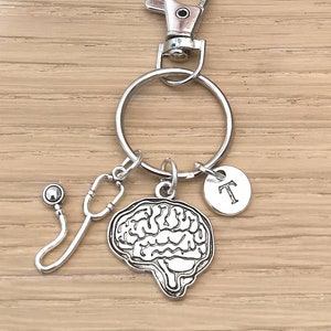 Può includere: Portachiavi d'argento con un ciondolo a forma di cervello, un ciondolo a forma di stetoscopio, un piccolo ciondolo rotondo con la lettera T e un anello diviso in argento.