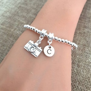 Peut inclure: Bracelet en argent avec des perles et des breloques. Il y a un appareil photo miniature et un médaillon rond avec la lettre "C". Le bracelet est porté au poignet.