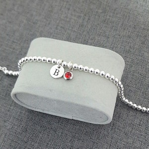 May include: A silver beaded bracelet with a silver charm featuring the letter "B" and a red gemstone charm.