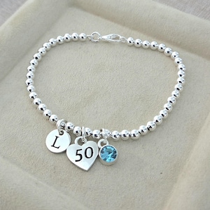 May include: Silver beaded bracelet with three charms. Charms include a heart with the number 50, a round charm with the letter "L", and a blue crystal. The bracelet is displayed on a beige surface.