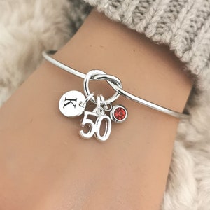 May include: Silver bangle bracelet with a knot detail and charms. The charms include a round charm with the letter "K" and a charm with the number "50". A small red gemstone is also attached to the bracelet.