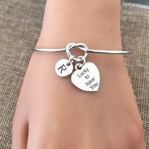 May include: Silver knot bangle bracelet with a heart charm that says "Lucky to have you" and a round charm with the letter "R".