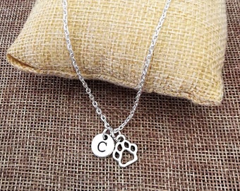 May include: A silver chain necklace with a small silver charm featuring a paw print and a letter 'C'. The necklace is displayed on a brown burlap surface.
