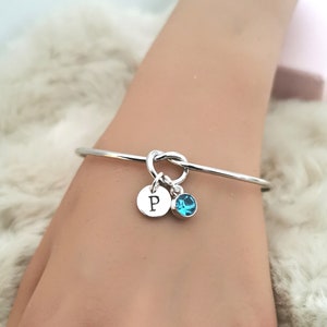 May include: A silver bangle bracelet with a knot design and two charms. One charm is a round disc with the letter "P" engraved on it. The other charm is a small, round, blue gemstone.