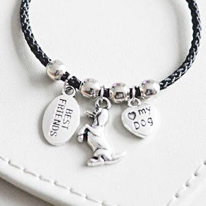 Puede incluir: Pulsera trenzada negra con dijes plateados. Los dijes incluyen un corazón con el texto "my dog", una silueta de perro y una etiqueta con el texto "best friends".