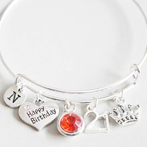 May include: Silver-toned bangle bracelet with multiple charms. Charms include a heart with "Happy Birthday" text, a red gemstone, the number "21", a crown, and a disc with the letter "N". The bracelet is on a white surface.