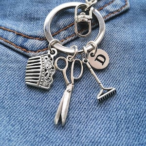 May include: Silver keyring with a comb, scissors, rake, and a charm with the letter D. The keyring is on a blue denim background.