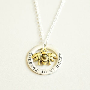 Puede incluir: Un collar de plata con un colgante circular que dice "forever in my heart". Un dije de abeja dorada cuelga del colgante.
